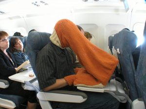 A man sitting on an airplane wearing a knit tube over his head and his laptop computer. His hands fit into little openings near the laptop.