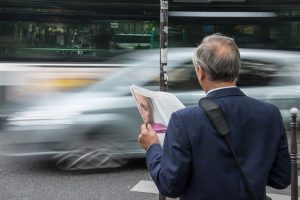 An older man holding a newspaper waiting for a car to pass. Different generations approach their business travel differently.