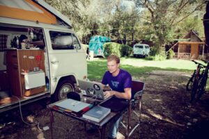 A man sitting at his computer outside, next to his van, living the life of a digital nomad.