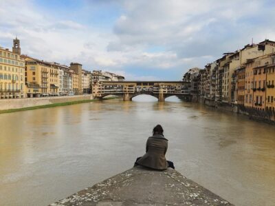 Woman sitting on a bank looking out over a river in Italy. This is something you can do on a solo travel trip.